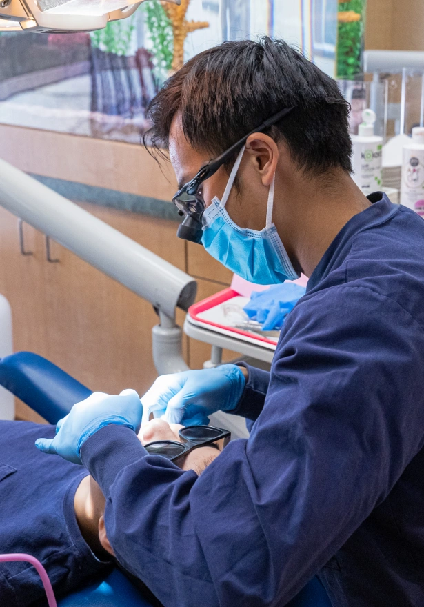 A male dental provider wearing a blue surgical face mask, protective glasses, and blue gloves performs a procedure on a patient in a dental chair.