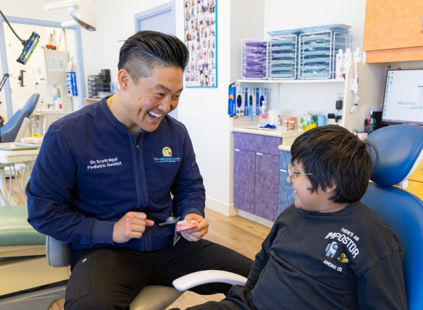 Dr. Scott Ngai smiles warmly while showing a dental model to a young patient sitting in a blue dental chair inside a brightly lit examination room.