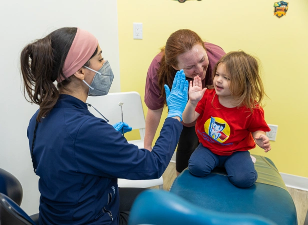 A pediatric dental provider wearing a face mask and blue gloves gives a high-five to a young child sitting on an examination table, while a parent smiles and watches in a brightly lit dental office.