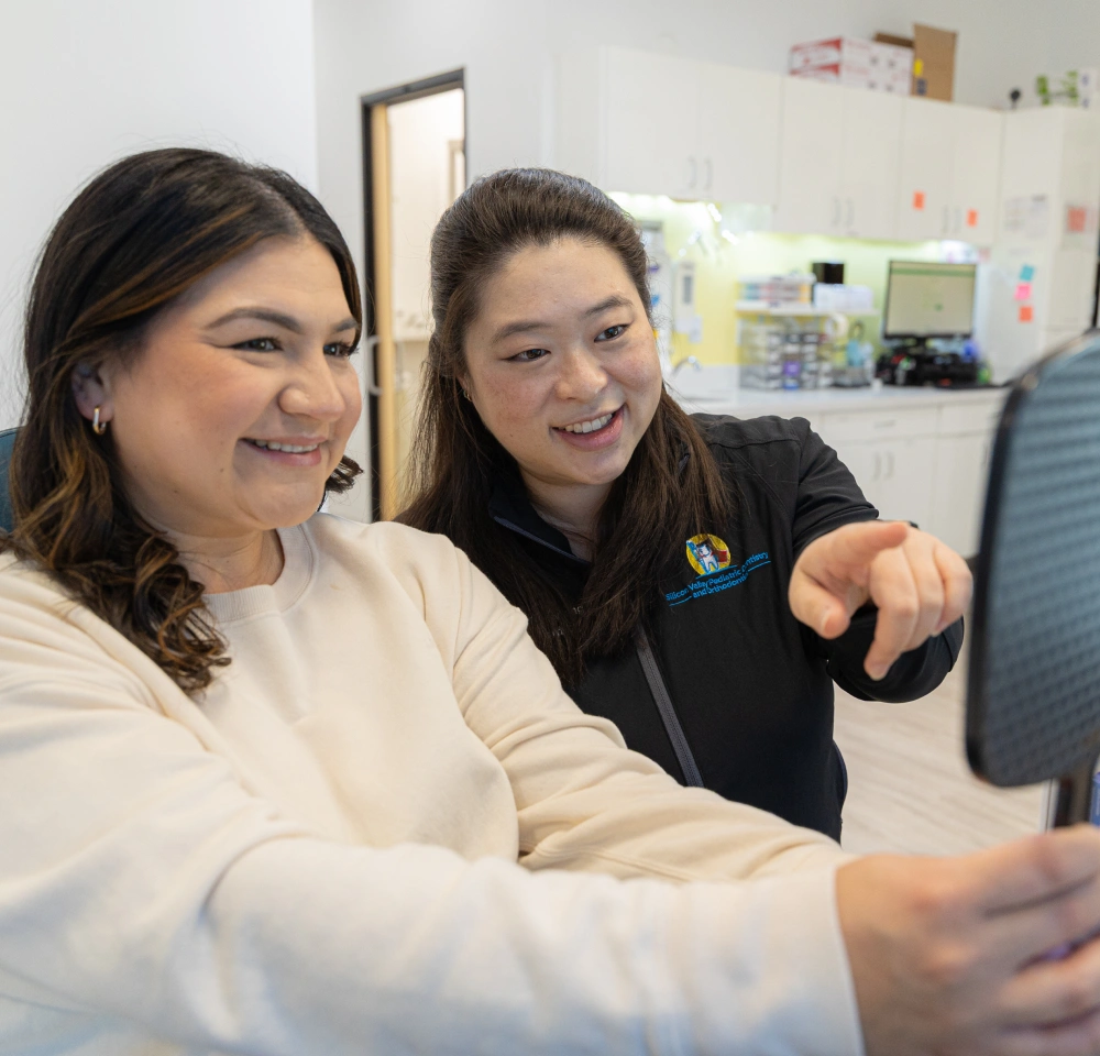 A smiling female dental staff member pointing out information to a seated patient in a clinical office setting.