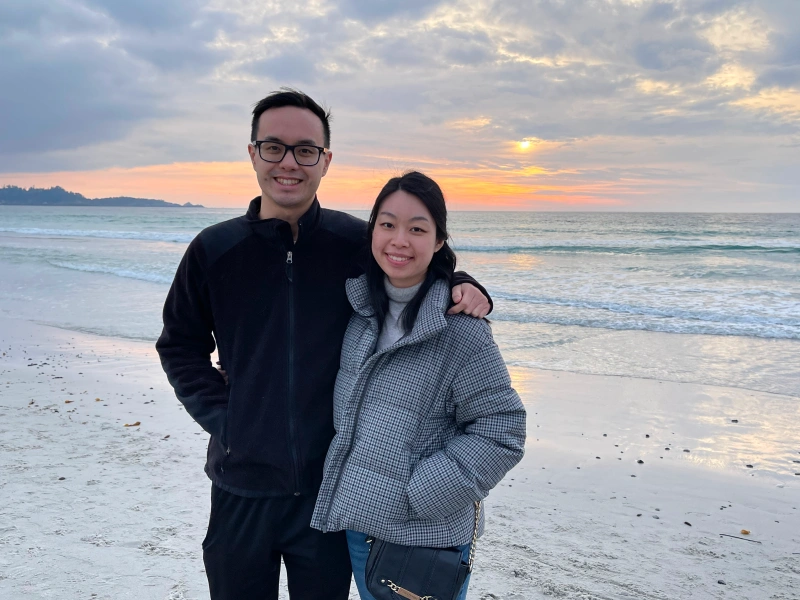 A smiling man and woman stand close together on a sandy beach during a vibrant sunset over the ocean.