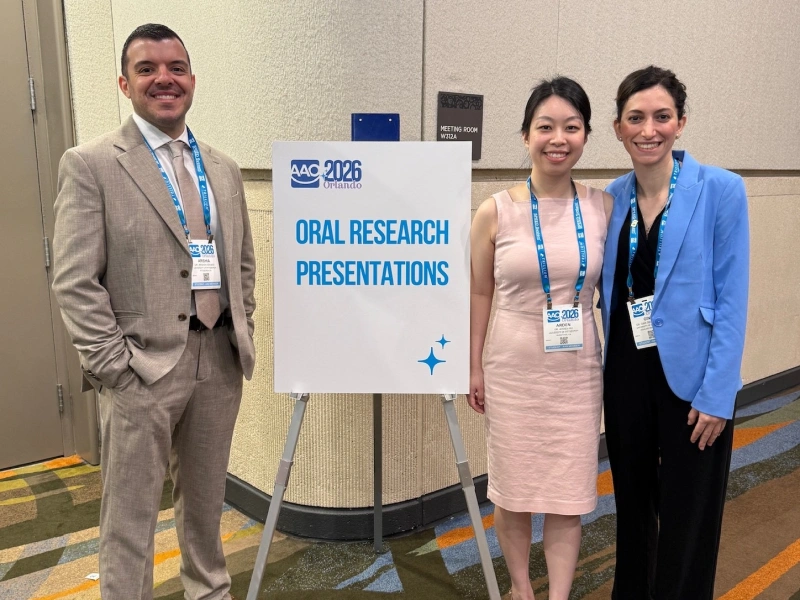 Three smiling professionals stand next to a sign that reads "Oral Research Presentations" at a conference.
