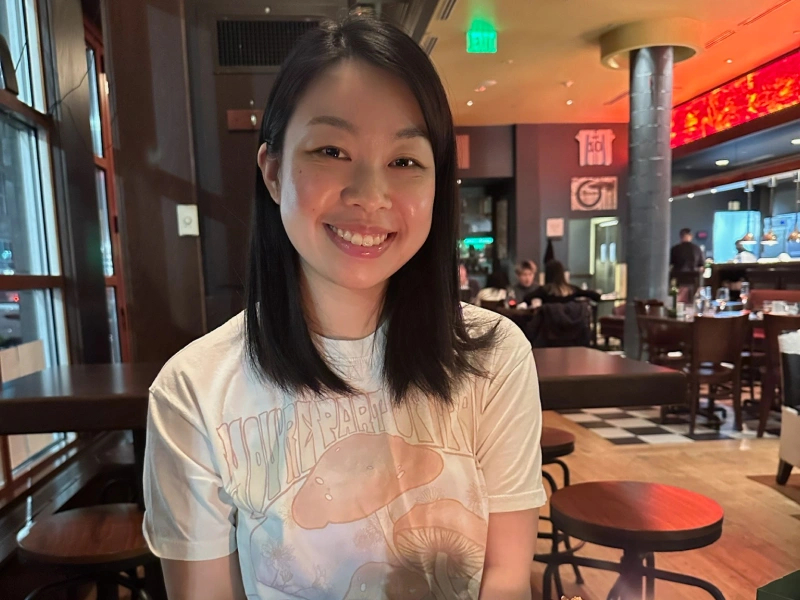 A smiling East Asian woman with shoulder-length straight dark hair, wearing a white graphic t-shirt with a mushroom design, seated inside a softly lit restaurant.