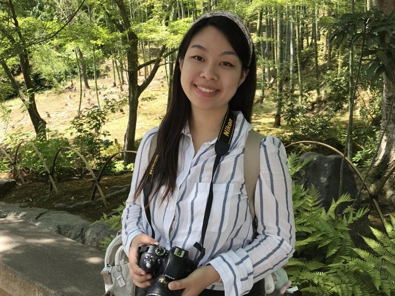 A smiling young woman holds a DSLR camera around her neck while standing on a path in a lush, green bamboo forest.