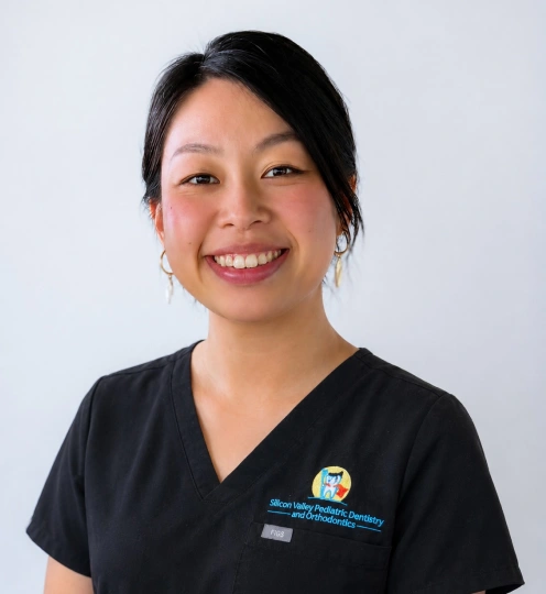 A headshot of a smiling woman with her black hair styled up and wearing thin, dangling hoop earrings. She is wearing black medical scrubs featuring a logo on the left chest pocket that reads "Silicon Valley Pediatric Dentistry and Orthodontics", set against a soft, light gray background.