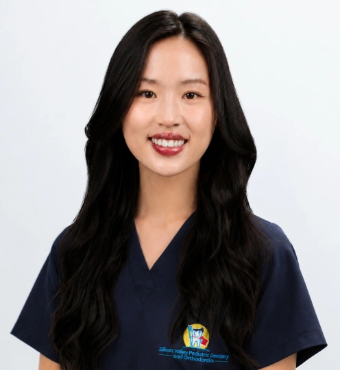 A headshot of a smiling woman with long, wavy black hair looking directly forward. She is wearing dark navy blue medical scrubs featuring a colorful logo on the left chest that reads "Silicon Valley Pediatric Dentistry and Orthodontics", set against a bright, plain white background.