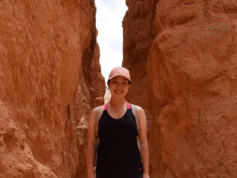 A smiling young woman wearing a pink baseball cap and black tank top stands inside a narrow red rock canyon.