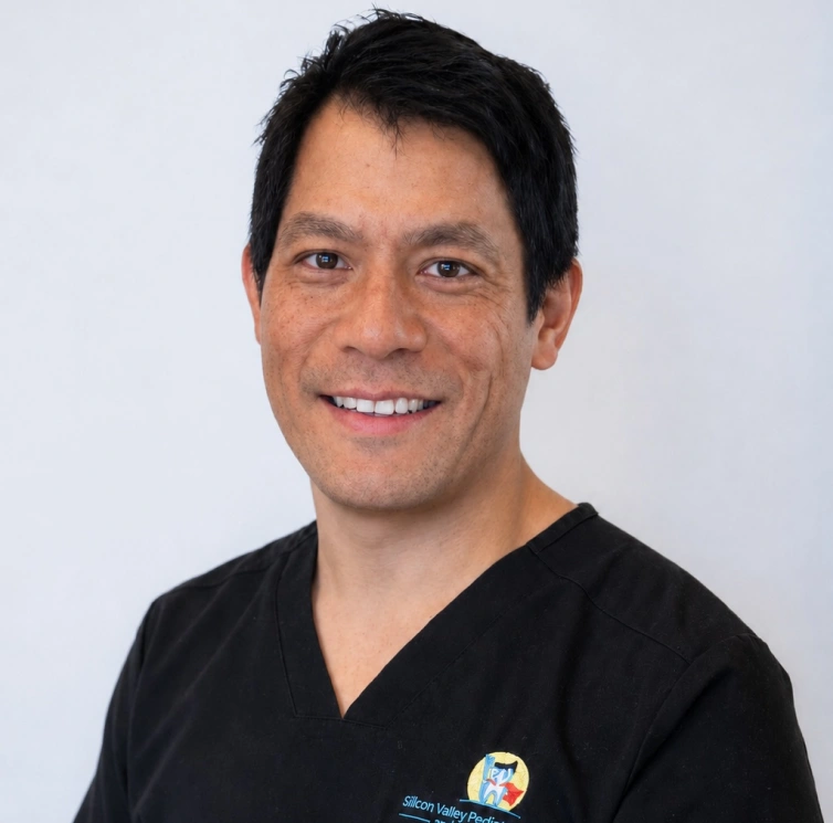 A headshot of a smiling man with short black hair and brown eyes, looking directly at the camera against a plain white background. He is wearing black medical scrubs featuring a small blue and yellow logo on the left chest that reads "Silicon Valley Pediatric".