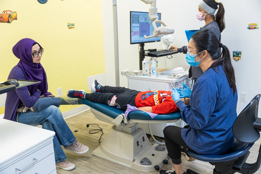 A dentist is examining a young child lying on a dental chair while the mother watches attentively from the side.